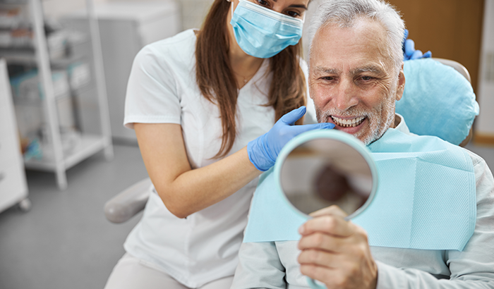 Older man in a dentist chair holding up a hand mirror and looking at his new dental implants at Periodontal Surgical Arts in Austin, TX