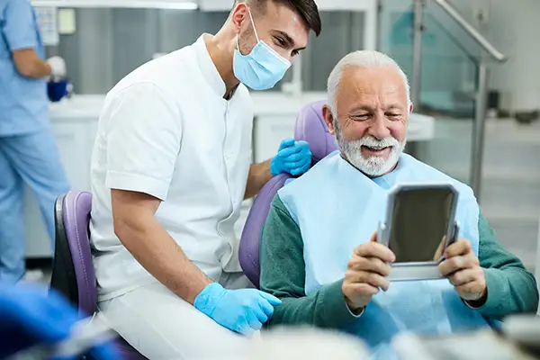 Happy, senior patient looking at himself in a mirror with dental implants at Periodontal Surgical Arts in Austin, TX